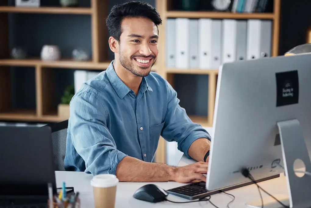 A man in a blue shirt is smiling while typing on a desktop computer in an office.